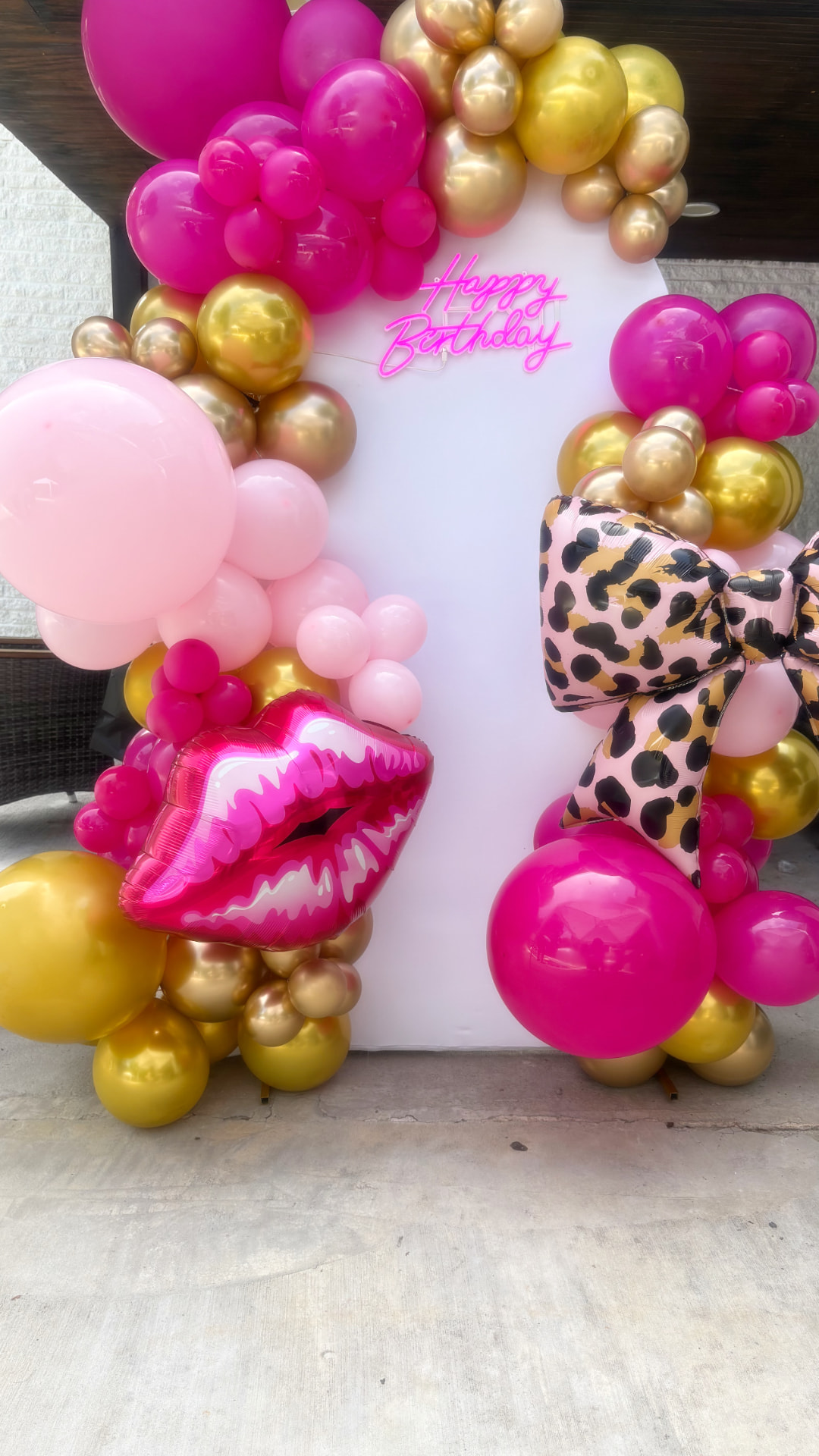White arch backdrop with hot pink Happy Birthday neon sign framed by a pink and gold balloon garland with lips and leopard bow foil balloons